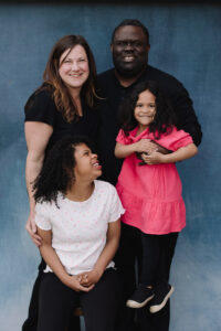 An image of a family including a white woman, a Black man, and two daughters. The younger daughter is being held up by the father and the other is sitting in front of the mother looking up at her sister and smiling.
